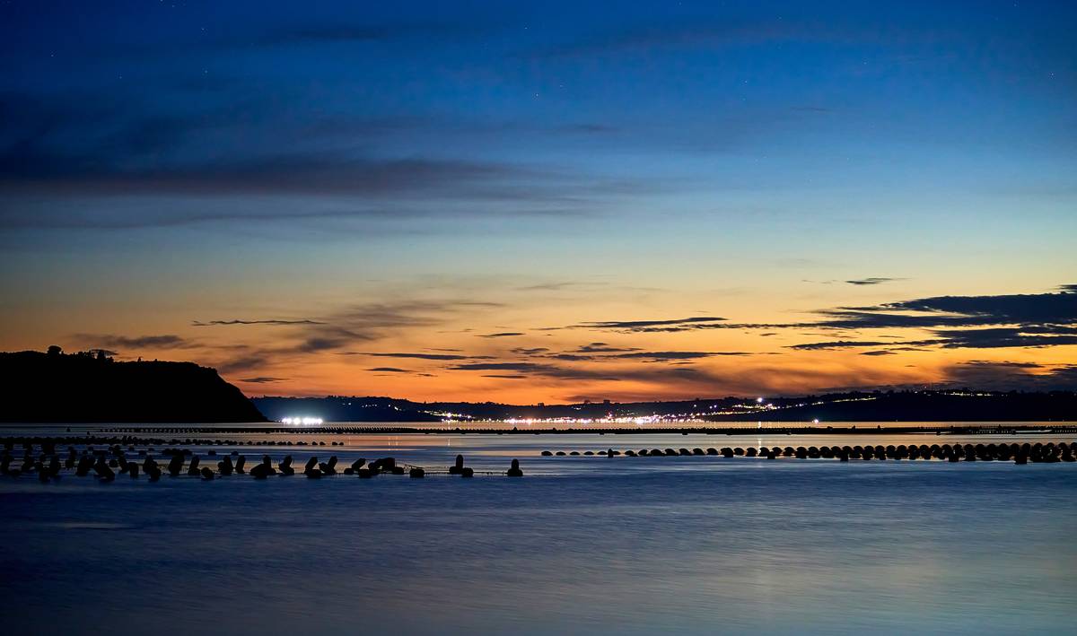 Peaceful Sunset at Calen Beach/Atardecer desde Playa Calen hacia Achao. A breathtaking sunset in Calen, Chiloé, Chile. The calm sea reflects shades of blue and orange, with mussel farming structures in the foreground. In the distance, the illuminated town and silhouetted hills complete the scene under a serene, partially cloudy sky. Un atardecer impresionante en Calen, Chiloé, Chile. El mar calmo refleja tonos azulados y anaranjados, con estructuras de cultivos de choritos en primer plano. A lo lejos, la ciudad iluminada y cerros oscuros en silueta completan la escena bajo un cielo sereno.